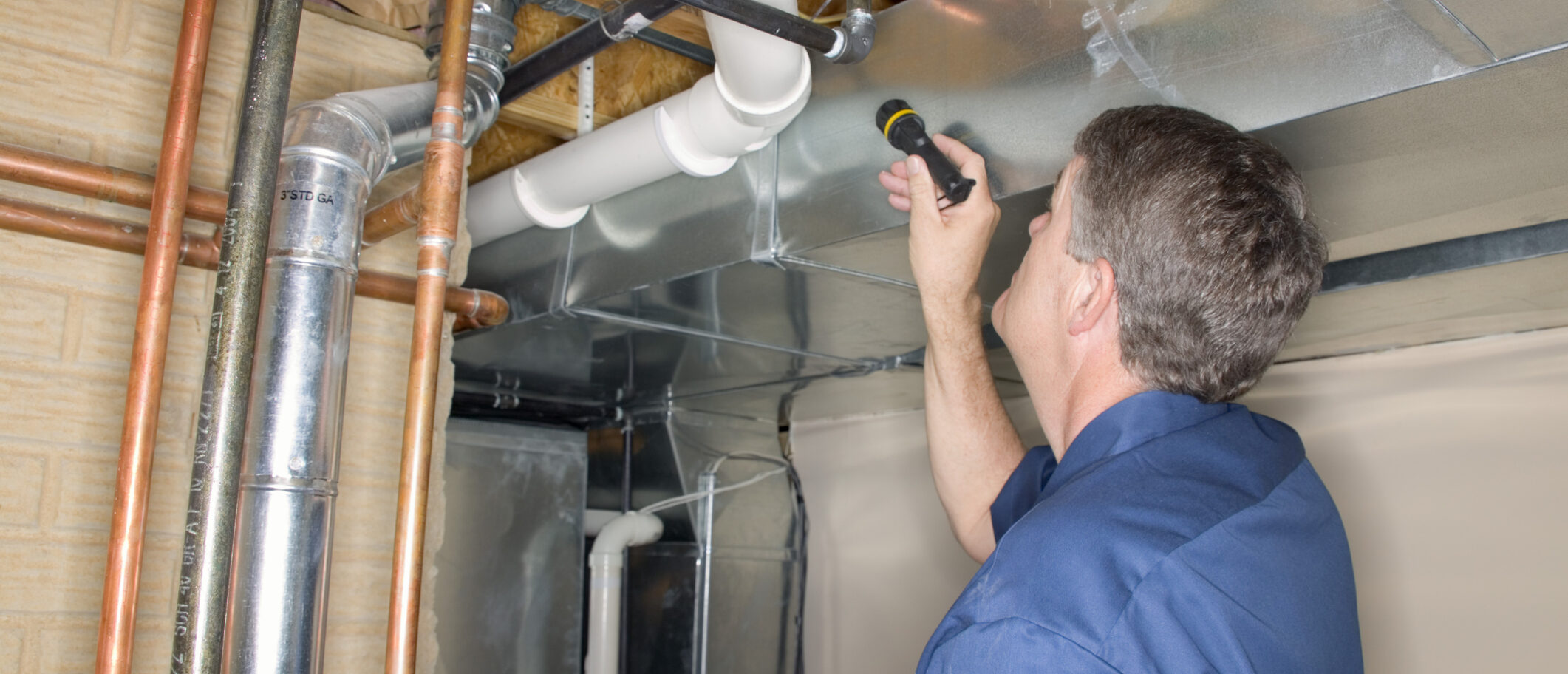 Man with a flash light evaluates basement plumbing during a home inspection.