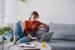 Woman in red shirt on couch transferring money via her laptop.