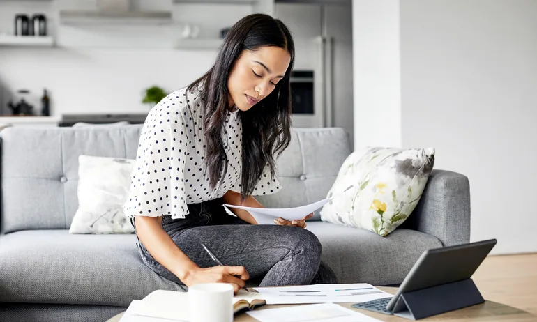 A woman sitting in a living room with papers and a tablet — an image of what learning how to invest money can look like.