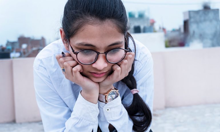 A female student, wearing round black-frame glasses and a white shirt, rests her chin in her hands