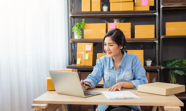 Young woman with her dark hair pulled back works at a laptop with paperwork on her desk.