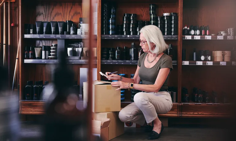 Senior saleswoman holding paper while crouching by boxes in deli