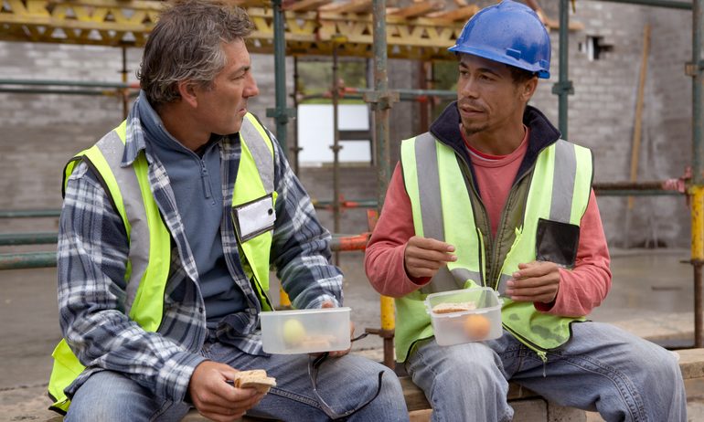 Two construction workers sitting down on a residential building site while eating their lunch.