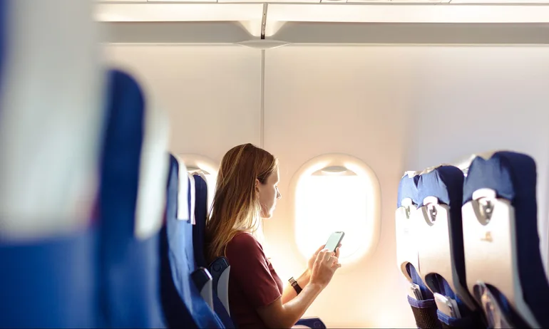 Young woman sitting on the aircraft seat near the window during the flight in the airplane. She is using smart phone with a blank screen