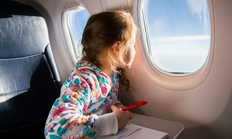 Child drawing picture with crayons in airplane. Little girl occupied while flying in aircraft. Travel with family and kids. Blue sky and sun outside the window