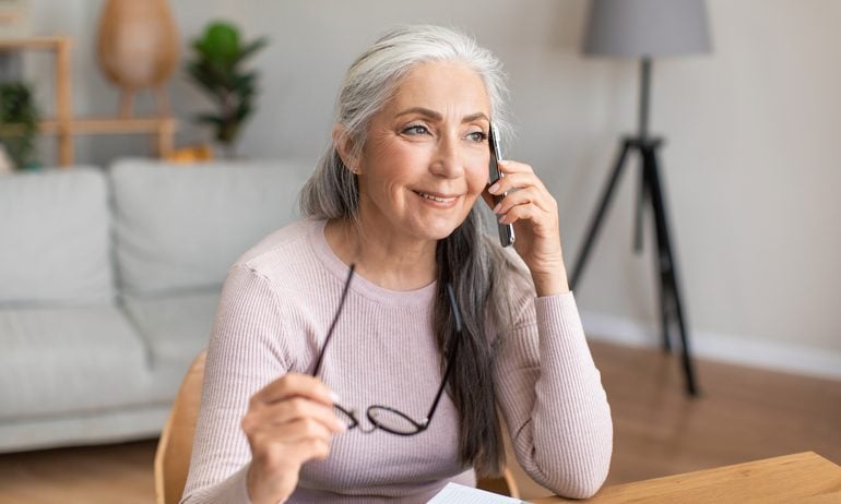 Smiling caucasian mature gray-haired lady hold glasses speaks by phone with client in living room interior