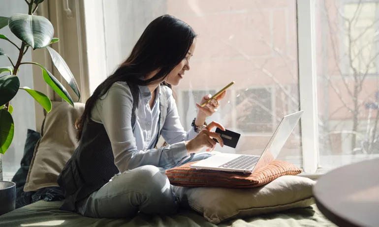 Woman with long dark hair sitting on a bed or couch next to a window looking at her phone to illustrate Holiday Deals Are Here. Don't Let Debt Follow